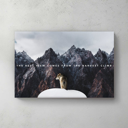 A motivational metal wall art print of a lone wolf on a snowy peak, surrounded by towering rugged mountains, with the text "The best view comes from the hardest climb."