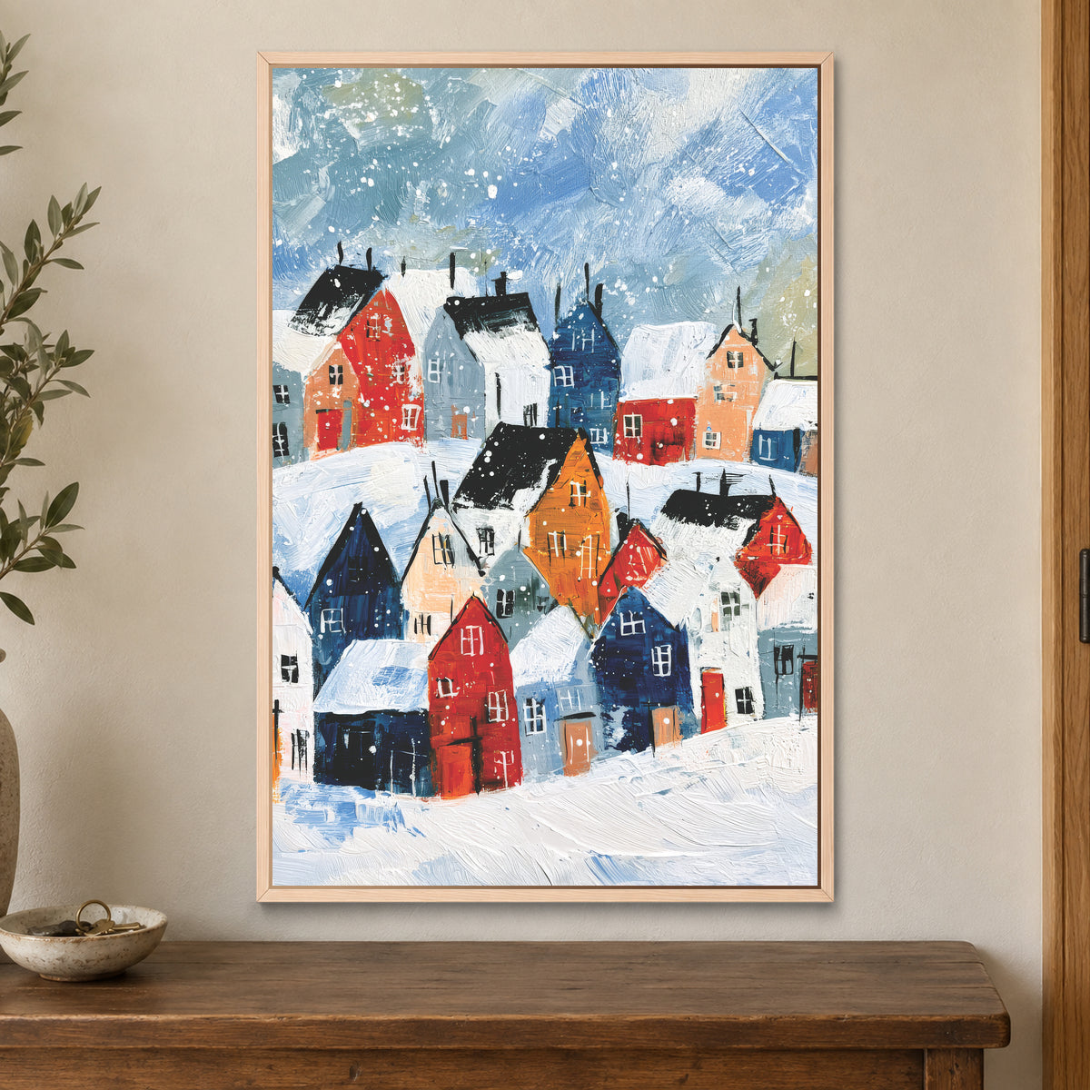 Snowfall Over the Rooftops framed canvas in a natural wood floating frame on a cream wall above a wooden sideboard beside a door frame