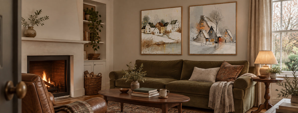 Two winter landscape canvas prints in oak frames above a green velvet sofa beside a glowing fireplace.