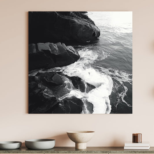 Black and white image of ocean waves splashing against rugged rocks along the shoreline.