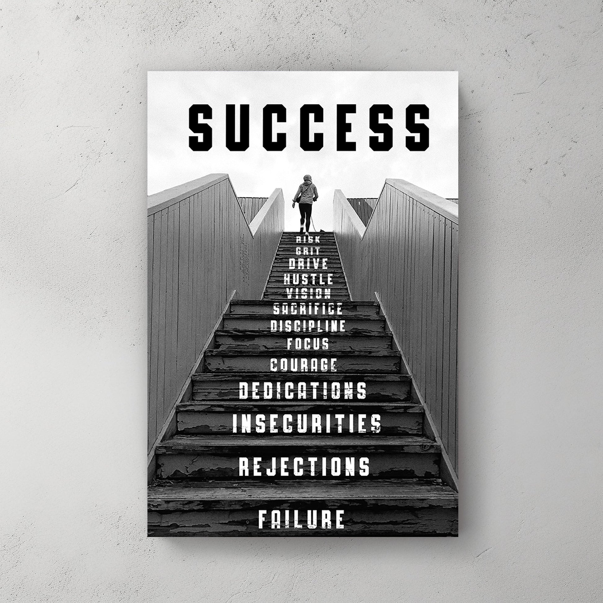 Black-and-white metal wall art print of a person climbing a staircase with motivational words like "risk," "focus," and "dedication" inscribed on the steps, representing the journey to success.