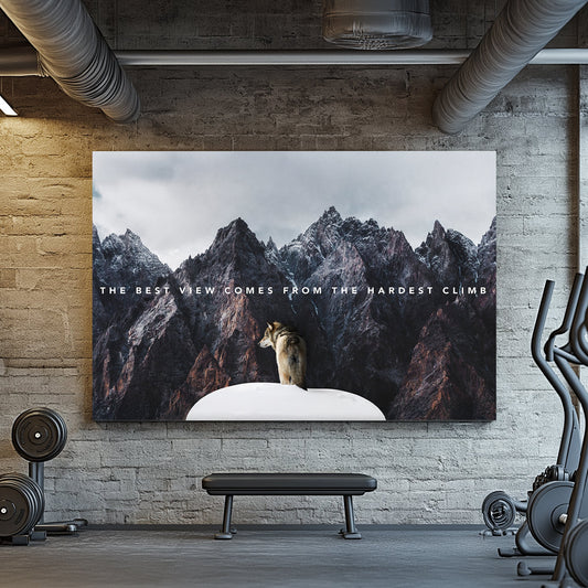 A motivational metal wall art print of a lone wolf on a snowy peak, surrounded by towering rugged mountains, with the text "The best view comes from the hardest climb."