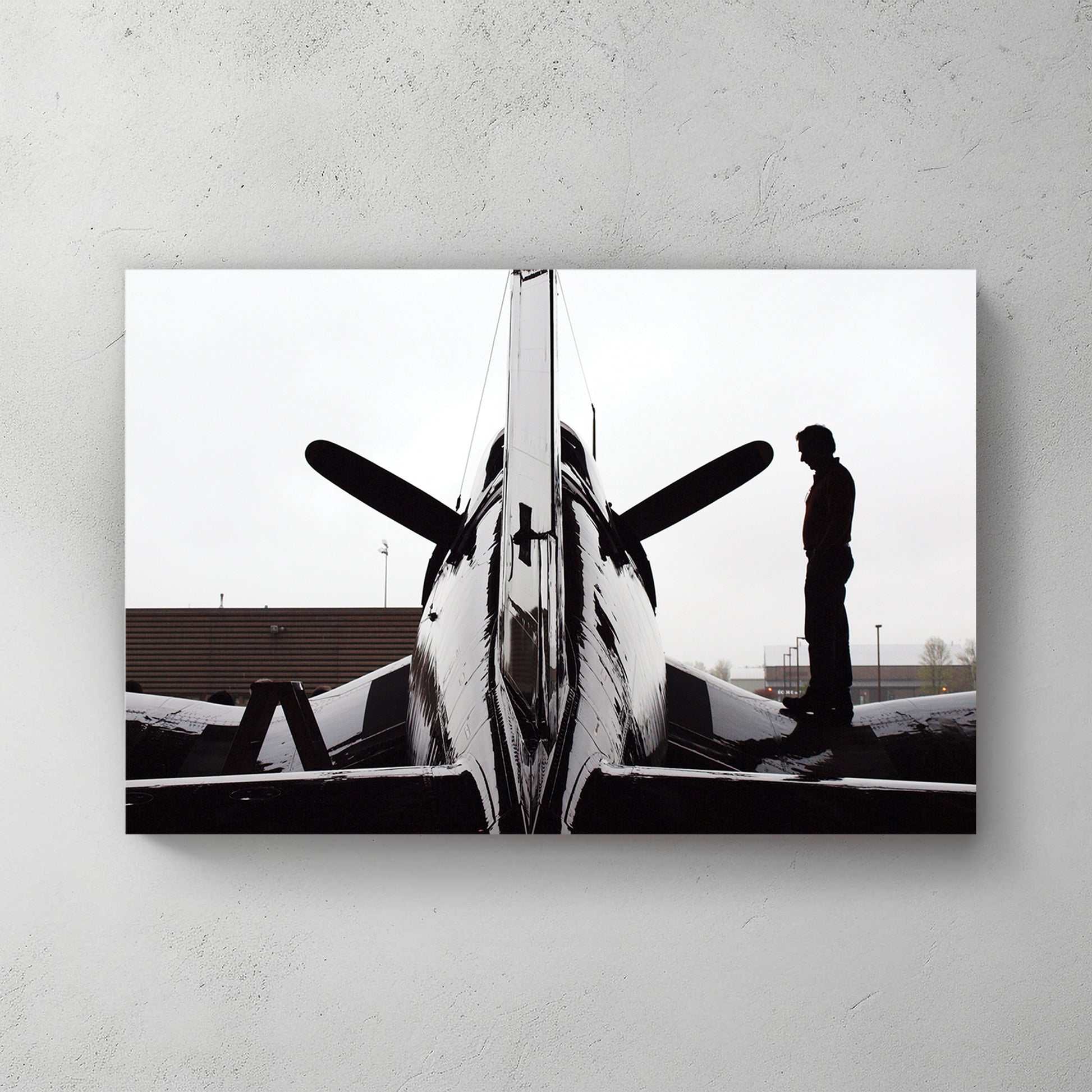 Silhouette of a man beside a WWII Corsair fighter plane in a hangar doorway, backlit grey sky — canvas wall art print