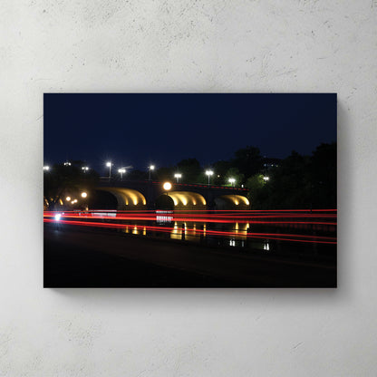 Night long-exposure of a lit stone arch bridge with red car-light trails streaking over water — canvas wall art print