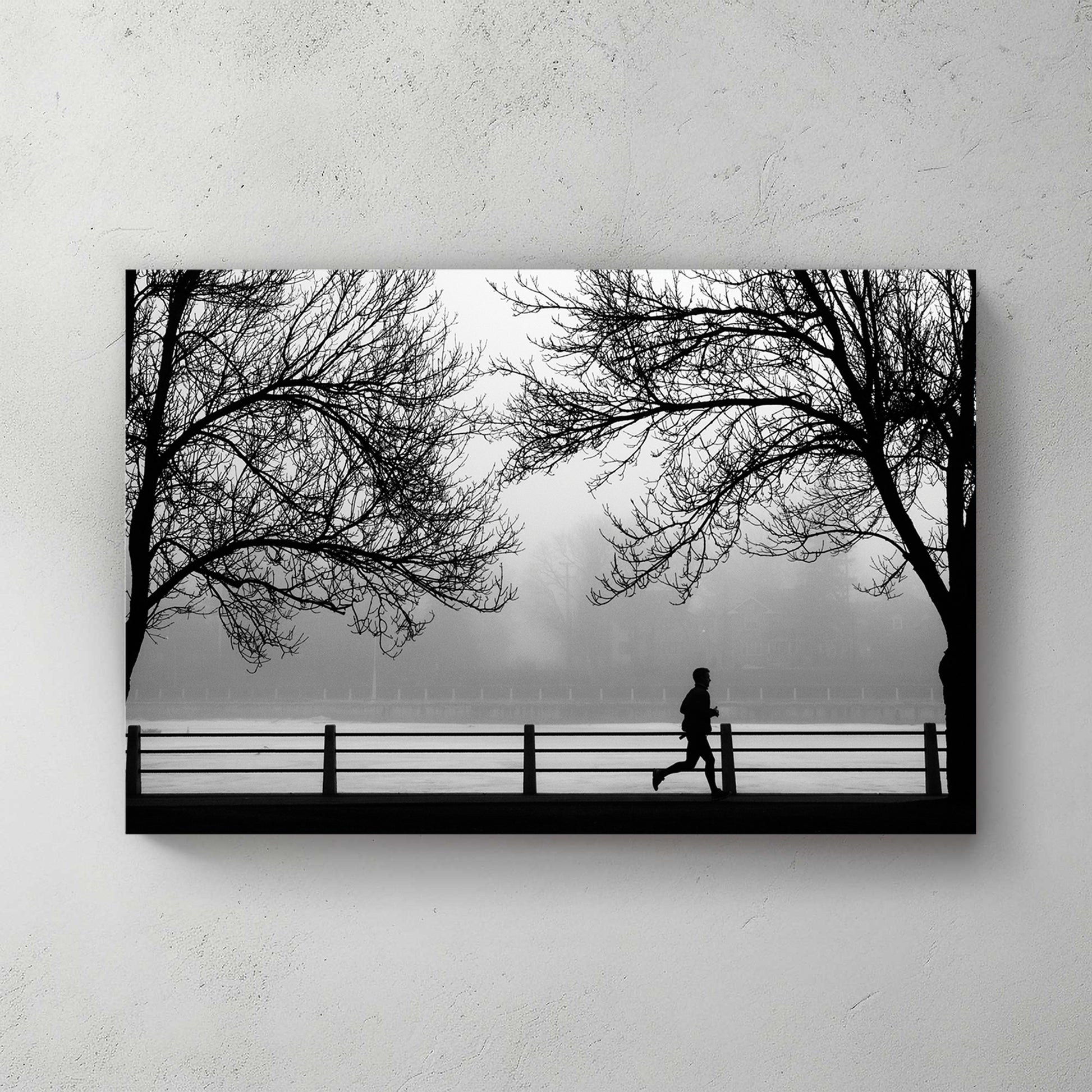 Silhouette of a lone jogger between two bare trees along a misty riverfront railing, moody grey — canvas wall art print