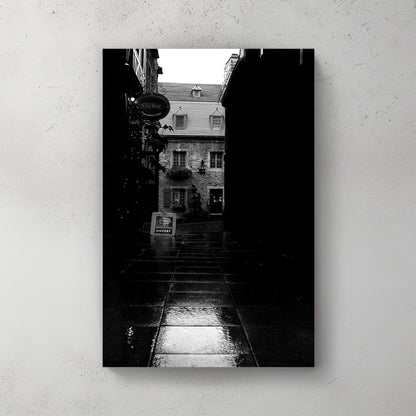 Moody black-and-white wet cobblestone alley in Old Quebec, a lit stone shopfront glowing at the end — canvas wall art print