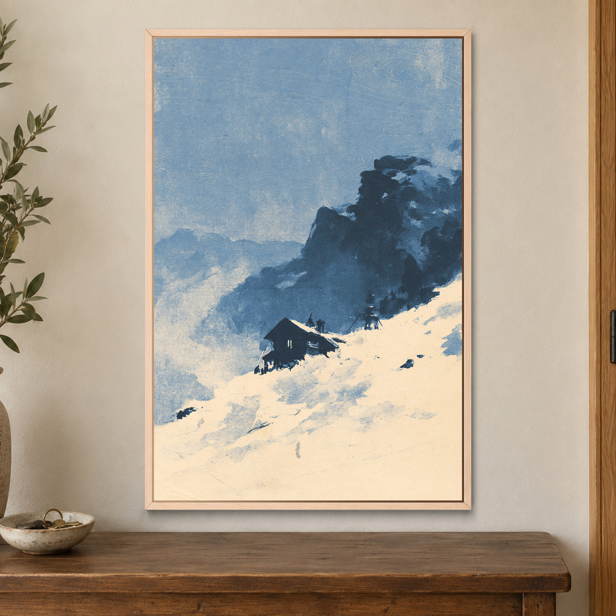 Lone Cabin Snow Slope framed canvas in a natural wood floating frame on a cream wall above a wooden sideboard beside a door frame